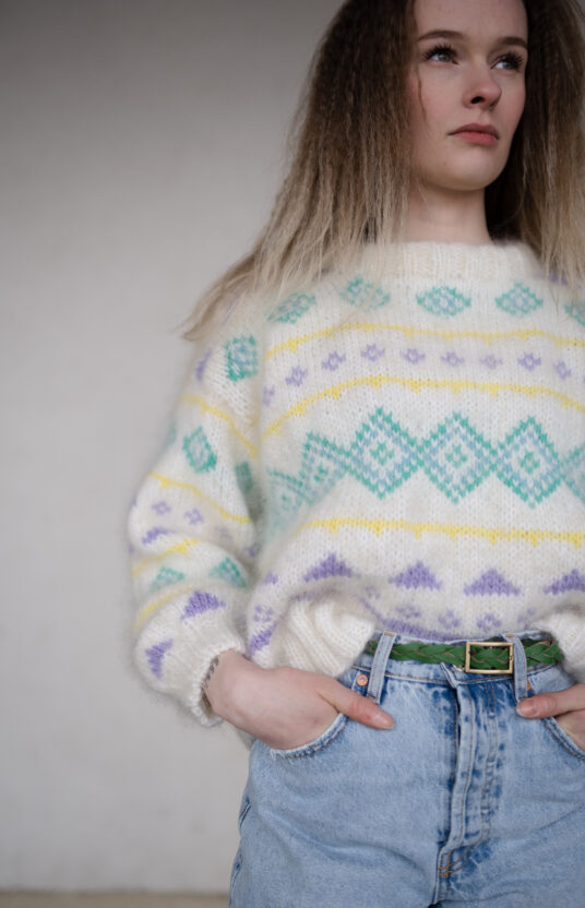 Person wearing a cream knit sweater with pastel diamond patterns and a green braided belt tucked into light-blue jeans, hands in pockets.