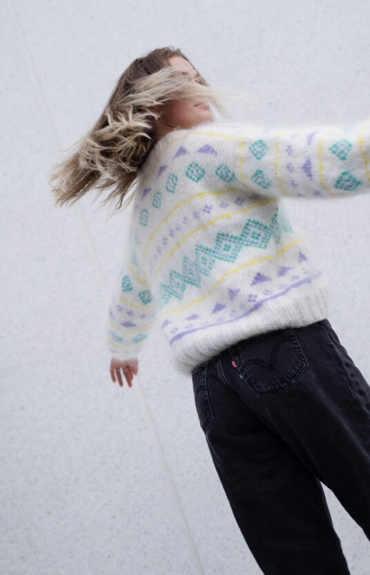 Person in a white patterned sweater with pastel shapes, arms stretched out, facing away against a light gray wall.