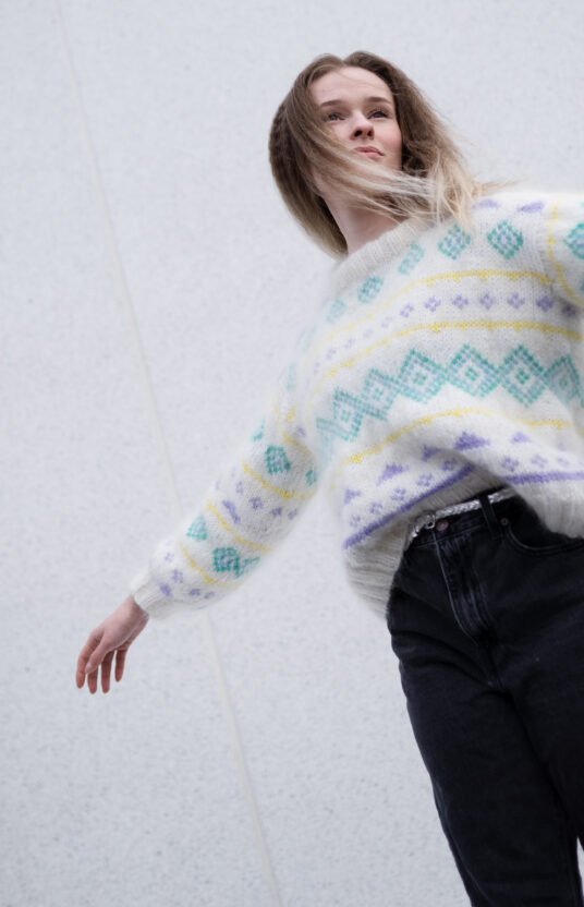 Person in a cream knit sweater with geometric teal and purple patterns, arms stretched outward against a pale gray wall.