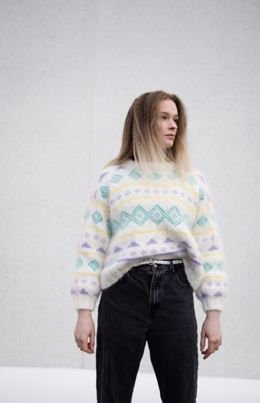 Person standing against a light gray wall, wearing a cream knit sweater with pastel diamond patterns and black jeans.